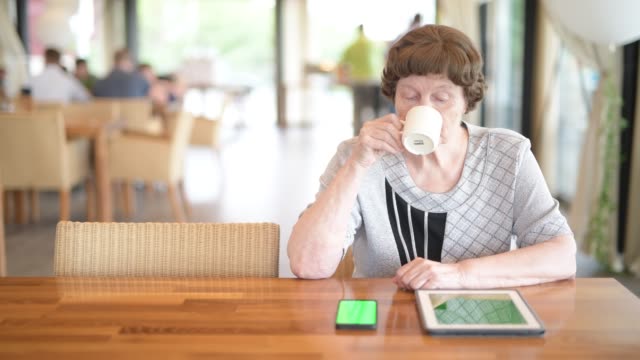 Senior-Frau-mit-Telefon-und-digitale-Tablet-beim-Kaffeetrinken