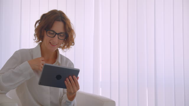 Closeup-portrait-of-adult-redhead-attractive-businesswoman-in-glasses-using-the-tablet-looking-at-camera-smiling-happily-sitting-in-the-armchair-in-the-white-office