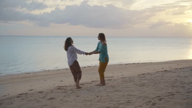 Zwei-junge-glückliche-Frauen-tanzen-und-spaßigen-sich-am-Strand-bei-Sonnenuntergang.
