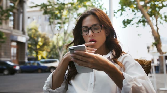 Business-female-in-glasses,-white-shirt.-She-feeling-disappointed-and-shocked-reading-news-using-cellphone,-sitting-in-roadside-cafe.-Slow-motion