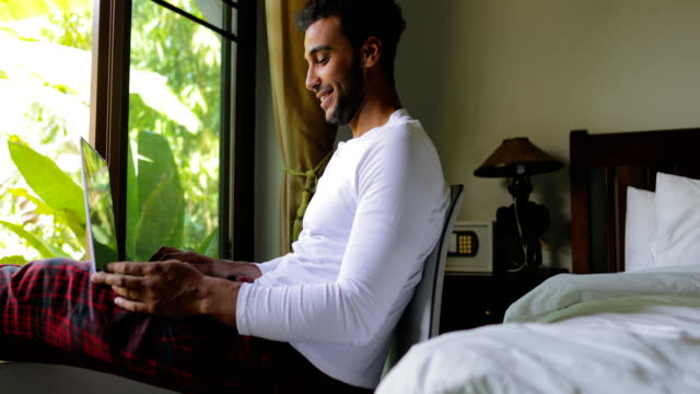 Hispanische-Jüngling-mit-Laptop-Computer-glücklich-lächelnden-Mann-im-Chat-Online,-über-große-Fenster-mit-Blick-auf-den-tropischen-Garten