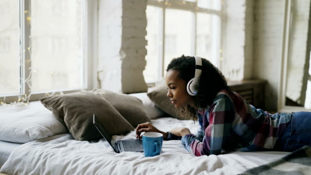 Chica-atractiva-raza-mixta-escuchar-música-mientras-que-los-medios-sociales-surf-en-laptop-acostado-en-cama
