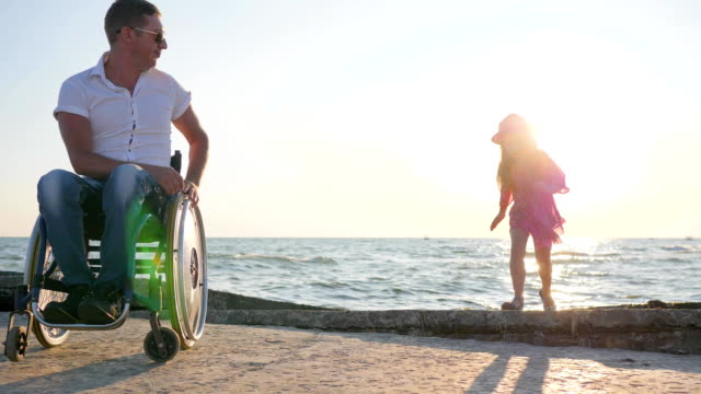 behinderte-Menschen-im-Rollstuhl-schaut-springen-Kind-bei-Gegenlicht-in-der-Nähe-von-Meer-in-Zeitlupe-applaudiert-Tochter