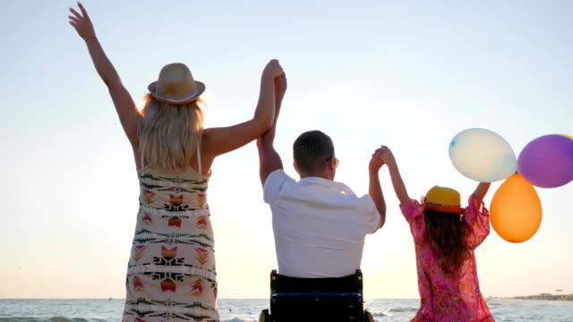 Invalid-with-wife-and-daughter-raise-their-hands-up-with-colorful-air-balloons-near-sea,-family-with-child