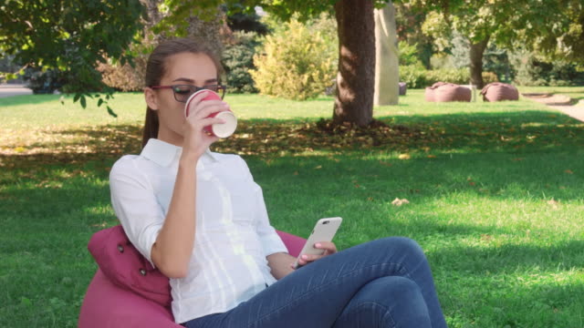 Young-woman-sitting-in-city-park-drink-coffee-and-browsing-on-phone