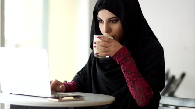 Joven-mujer-musulmana-en-hijab-sentada-en-el-café-bebiendo-una-taza-de-café-y-el-uso-de-su-portátil.-Ella-está-buscando-algo-en-internet.-Estudiando-o-trabajando.-Tiro-de-cámara-lenta
