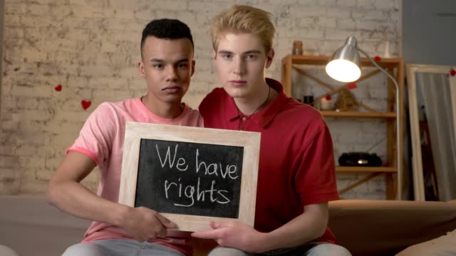 A-sad-international-gay-couple-is-sitting-on-the-couch-and-holding-a-sign.-We-have-rights.-Look-at-the-camera.-Home-comfort-on-the-background.-60-fps
