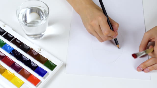Nicht-erkennbare-Frau-zieht-eine-Bleistiftskizze-auf-Papier-Osterkarte.-Auf-dem-Tisch-ist-eine-Palette-mit-Farben-und-ein-Glas-Wasser.
