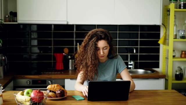 young-pretty-curly-caucasian-woman-sitting-alone-at-table-in-nice-spacious-kitchen-idoors-working-on-laptop-listening-to-music,-printing-on-laptop-and-dancing
