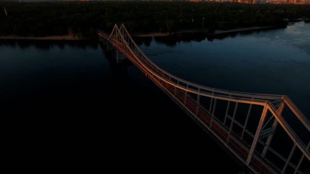 Pedestrian-bridge-over-the-river-near-the-city-at-sunset-aerial-drone-shoot