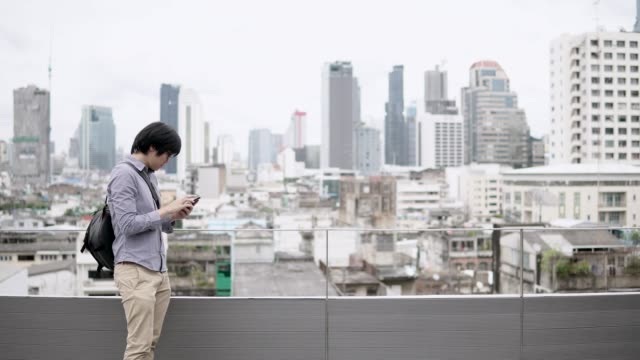Junge-asiatische-Geschäftsmann-mit-Smartphone-auf-Bürogebäude-Dachterrasse-mit-Blick-auf-die-Stadt-im-Hintergrund.-Verwendung-von-social-Media-Anwendung-oder-spielen-auf-Smartphone-Konzepte.