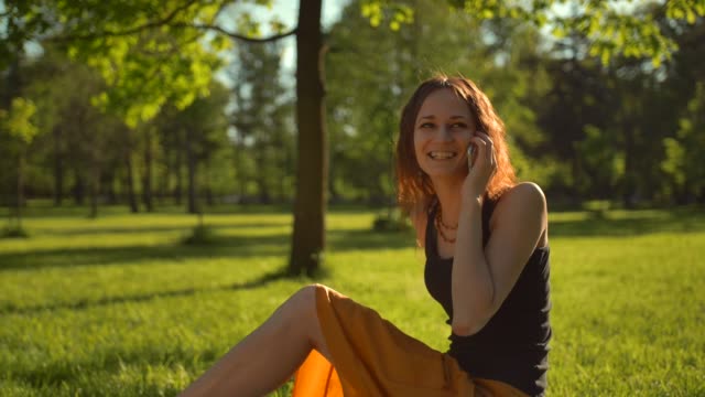 Glücklich-schön-weiblich-sitzend-auf-grünen-Rasen-und-haben-Gespräche-über-digitale-Smartphone-Gerät