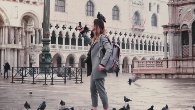 Happy-smiling-female-tourist-with-pigeons-sitting-on-her-arm-and-head-takes-selfie-on-city-square-in-Venice-slow-motion.
