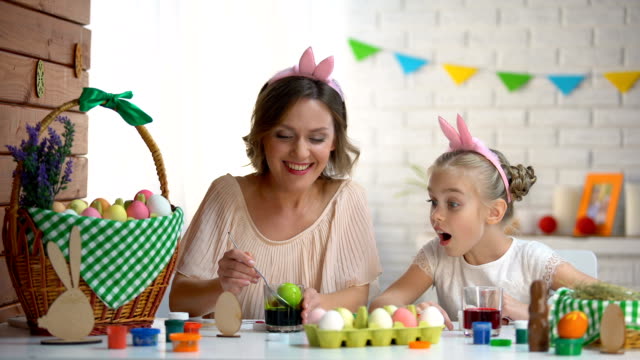Little-girl-excitedly-watching-mother-dipping-egg-in-green-food-coloring,-Easter