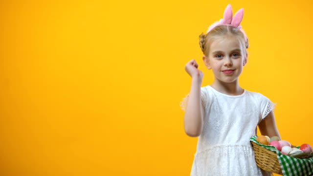 Female-child-in-bunny-ears-headband-holding-Easter-basket-and-showing-thumbs-up