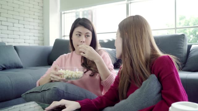 Asian-women-playing-pillow-fight-and-eating-popcorn-in-living-room-at-home,-group-of-roommate-friend-enjoy-funny-moment-while-lying-on-the-sofa.-Lifestyle-women-relax-at-home-concept.