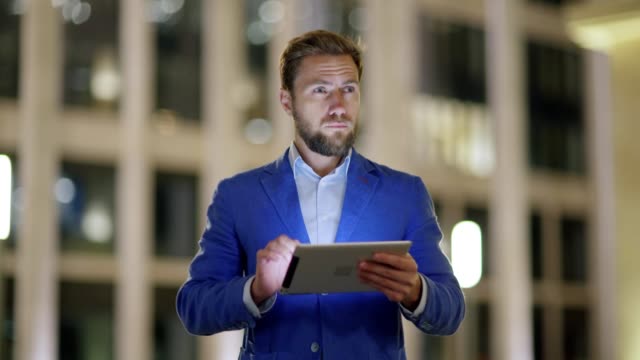 Medium-shot-of-handsome-bearded-middle-aged-man-in-blue-suit-walking-down-evening-street-and-browsing-net-on-tablet-pc