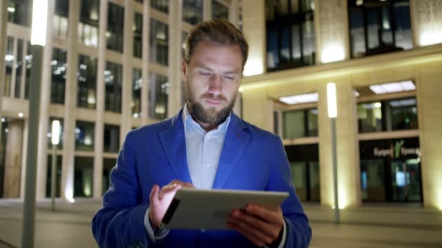 Medium-dolly-shot-of-handsome-middle-adult-businessman-surfing-Internet-on-tablet-computer-while-walking-outdoors-in-night-city-street