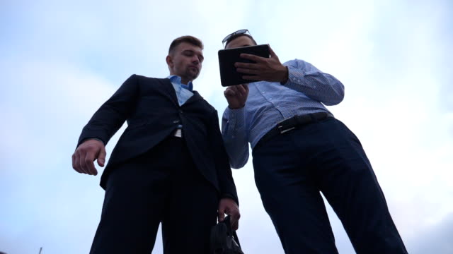 Low-angle-view-of-young-businessman-showing-a-presentation-on-screen-of-tablet-pc-to-his-colleague-in-city.-Successful-entrepreneurs-working-on-digital-tablet.-Blue-sky-at-background.-Slow-motion