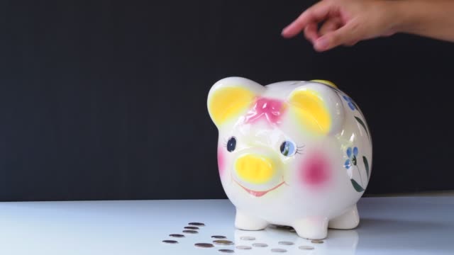 woman-hand-putting-coin-into-colorful-piggy-bank-white-table-and-black-background.-4k-b-roll-motion-footage
