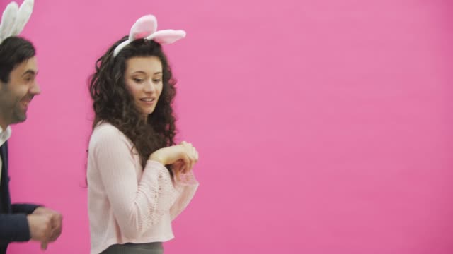 A-young-lovers-couple-appears-on-the-pink-background,-reproducing-horses-of-hares.-With-the-ears-of-a-pink-rabbit-on-the-head.-Easter-concept.-Slow-video.