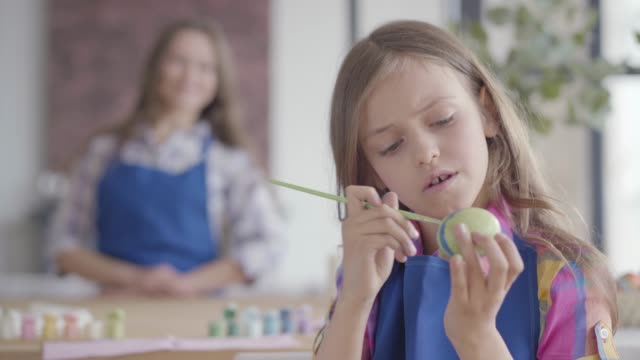 Portrait-of-cute-concentrated-girl-with-long-hair-in-blue-apron-painting-Easter-egg