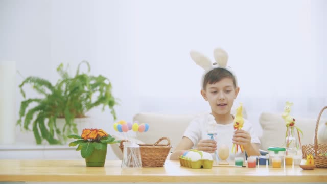 Joven-lindo-chico-está-sentado-en-la-mesa-llena-de-decoraciones-de-Pascua-y-está-jugando-con-conejitos-de-Pascua-en-sus-manos.-Teatro-conejito.