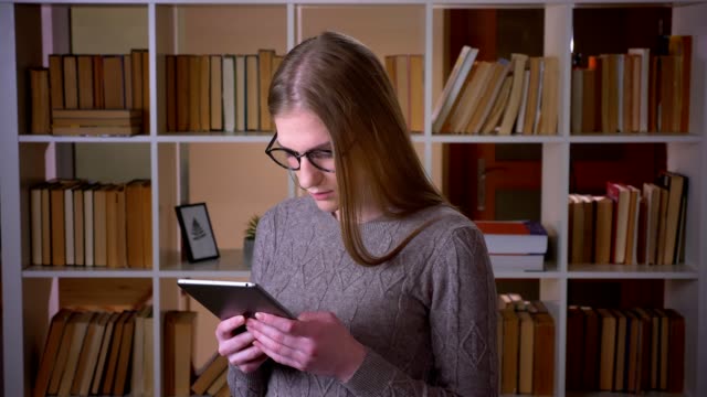 Retrato-de-primer-plano-de-joven-atractiva-estudiante-en-gafas-usando-la-tableta-y-sonriendo-mirando-la-cámara-en-la-biblioteca-de-la-universidad-en-el-interior