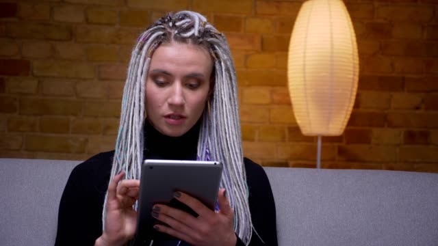 Closeup-shoot-of-young-attractive-caucasian-hipster-female-using-the-tablet-sitting-on-the-couch-indoors-in-a-cozy-apartment
