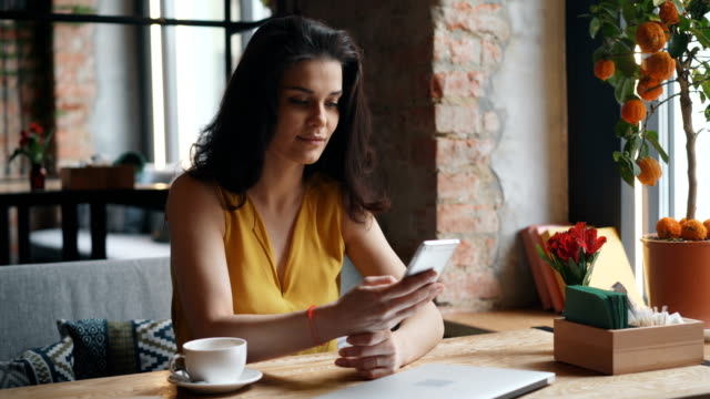 Charming-young-lady-holding-smartphone-touching-screen-smiling-in-modern-cafe