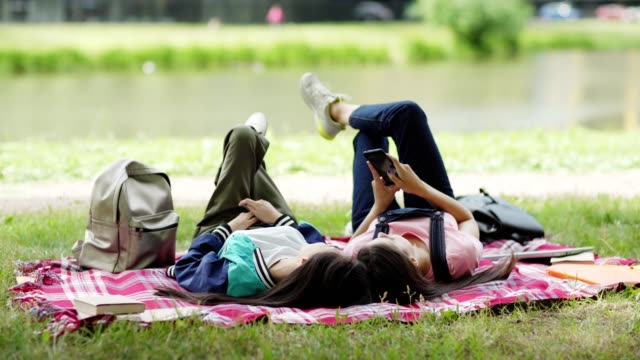 Two-college-friends-relaxing-on-lawn-near-pond-after-classes.-Students-lying-on-blanket,-browsing-smartphone-and-talking