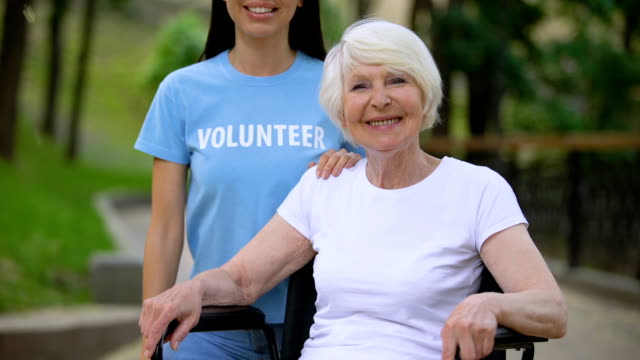 Mujer-madura-feliz-en-silla-de-ruedas-y-joven-voluntaria-buscando-cámara,-caridad