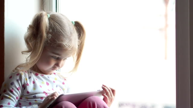 Blonde-little-girl-using-tablet-pc.-Sitting-on-the-window-sill.-Child-smiling-and-looking-at-camera.-Child-plays-on-the-PC