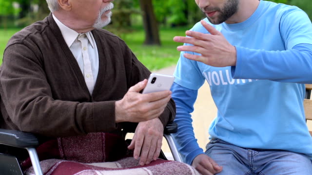 Male-volunteer-teaching-aged-man-in-wheelchair-how-to-use-smartphone,-caregiving