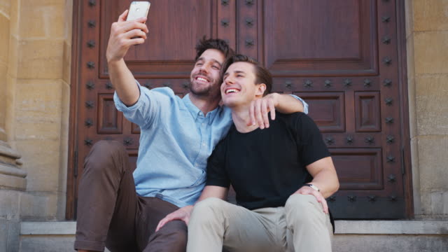 Männliche-Homosexuell-Paar,-das-Selfie-auf-handy-sitzt-im-Freien-auf-Stufen-des-Gebäudes