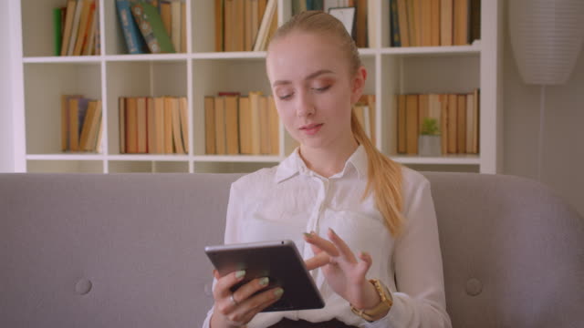 Nahaufnahme-Porträt-der-jungen-hübschen-kaukasischen-blonden-Studentin-mit-dem-Tablet,-das-auf-die-Kamera-schaut-und-glücklich-auf-der-Couch-in-der-Wohnung-sitzt