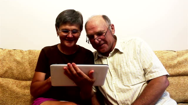 Una-pareja-de-ancianos-está-sentada-en-un-sofá-en-casa-y-viendo-fotos-en-una-tableta-PC,-sonriendo-y-hablando-activamente.