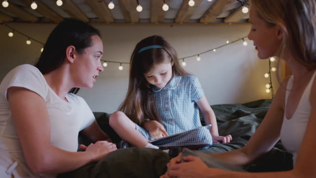 GleichGeschlecht-weibliche-paar-sitzen-auf-dem-Bett-mit-Tochter-zu-Hause-zusammen-mit-digitalen-Tablet
