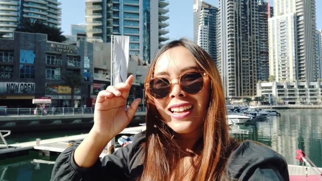 Cheerful-young-woman-takes-selfie-portrait-using-smartphone-sharing-travel-friends-showing-at-Dubai-marinaUnited-Arab-Emirates,-Dubai.
