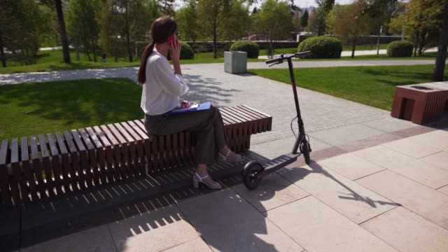 Businesswoman-talking-on-the-phone,-sitting-in-a-park-next-to-a-scooter.-Young-modern-woman-in-business-attire.-Conversation-outdoor
