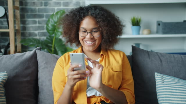 Schöne-afroamerikanische-Mädchen-mit-Smartphone-lachen-entspannend-zu-Hause