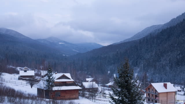 Timelapse-of-wooden-cottages-in-mountain-village-valley-surrounded-with-coniferous-forest-and-snowy-mountains.-Fast-view-of-chalets-covered-with-snow-at-ski-resort.-Day-to-night-evening-timelapse