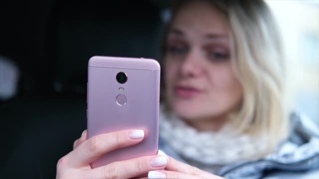 Verschwommene-blonde-Frau-Bloggerin-in-Winterkleidung-sitzen-im-Auto-und-im-Gespräch-mit-Anhängern,-Live-Streaming,-Blick-auf-Smartphone-Bildschirm.-Fokus-auf-Telefon
