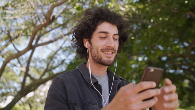 Portrait-of-a-smiling-young-man-with-earphones-in-his-ears-chatting-on-social-media-on-smartphone-in-the-park