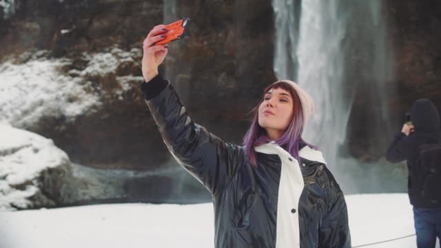 schöne-junge-Frau-in-der-Nähe-eines-Wasserfalls-in-Island,-macht-Fotos-am-Telefon,-freut-sich-und-macht-ein-Selfie