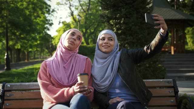 Two-muslim-women-in-stylish-hijabs.-Smiling,-taking-selfie-on-smartphone-and-enjoying-coffee.-Sitting-on-wooden-bench-in-park.-Сlose-up,-slow-motion