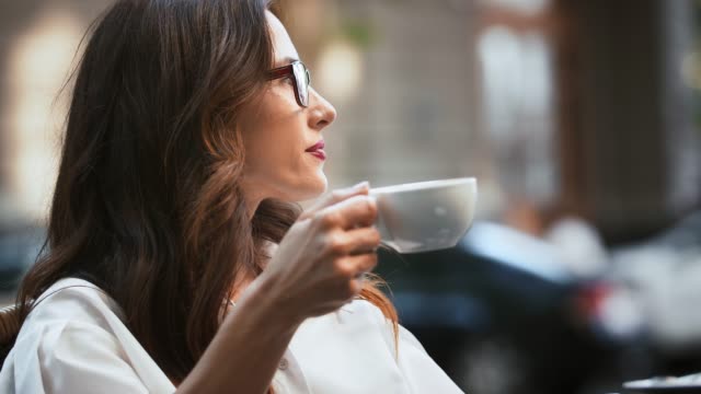 Business-lady-in-glasses-and-white-shirt.-She-sitting-at-table-in-outdoor-cafe.-Reading-news-on-cellphone-and-drinking-coffee.-Close-up