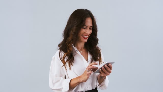 Brunette-woman-in-white-shirt-is-typing-on-smartphone,-smiling,-rejoicing-and-crossing-her-fingers-while-posing-on-gray-background.-Close-up