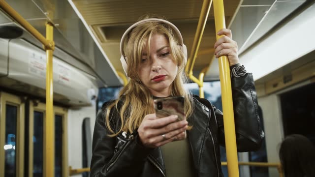 Young-tired-girl-hipster-in-headphones-is-listening-to-music.-Riding-tram,-using-smartphone,-standing-and-holding-onto-the-handrail.-Slow-motion