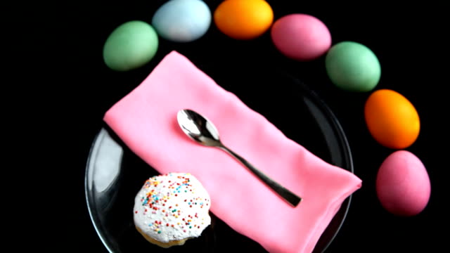 On-black-table-lay-different-colored-eggs,-Easter-cake,-plate-spoon,-and-napkin.